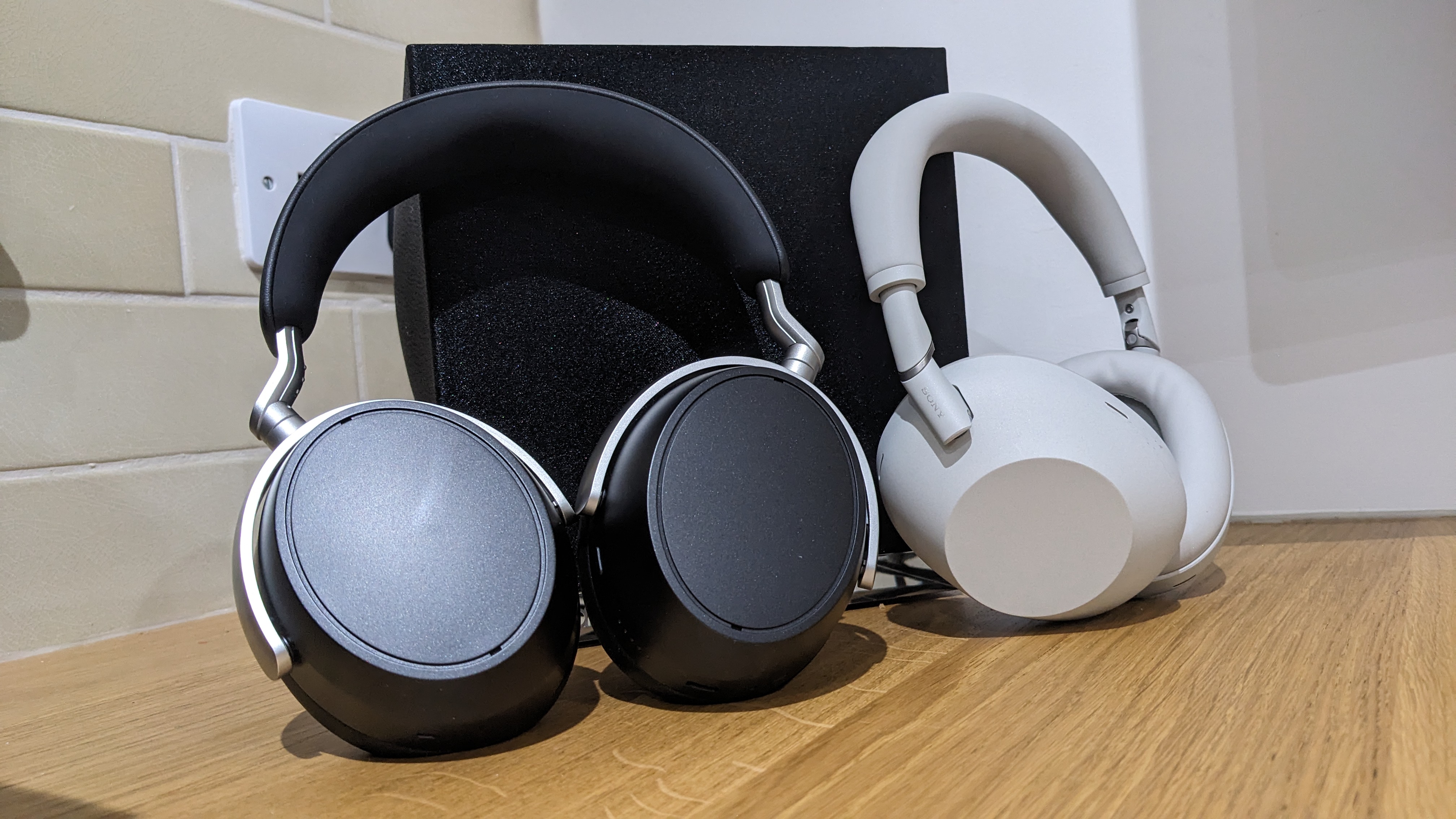A black pair of Sennheiser HDB 630 and white pair of Sony WH-1000XM6 over-ear headphones on a wooden surface.