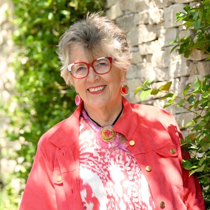 Prue Leith in coral jacket and blouse leaning against wall in garden