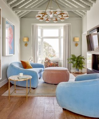living room with chunky, rounded furniture pieces, pale blue sofa and lounge chair, pale pink ottoman, metallic side table, dark wood flooring with rug, gray painted walls with high ceiling and painted beams, artwork, tv and fireplace on walls, golden wall lights and large pendant light