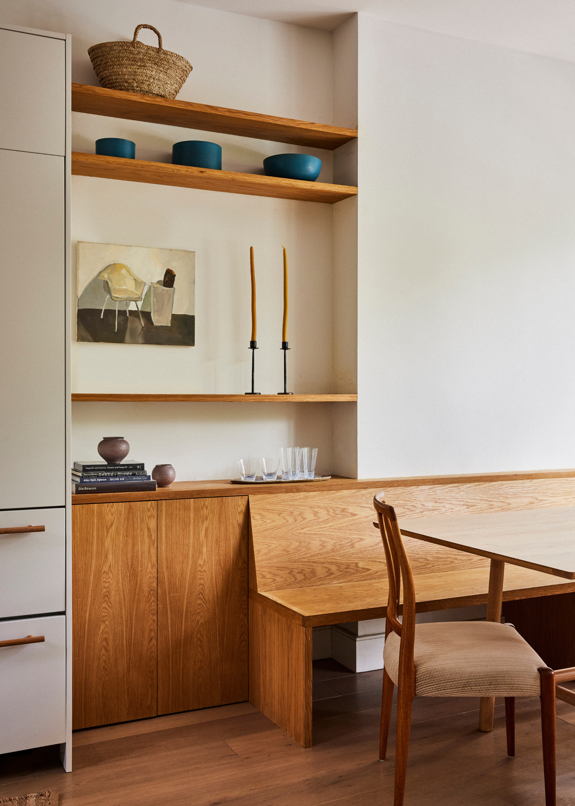A dining area with a wooden bench, a dining chair, a side storage space with wood shelves and cabinets featuring art, a set of bowls, a woven basket, a pair of candlesticks, a tray of glasses, and some books
