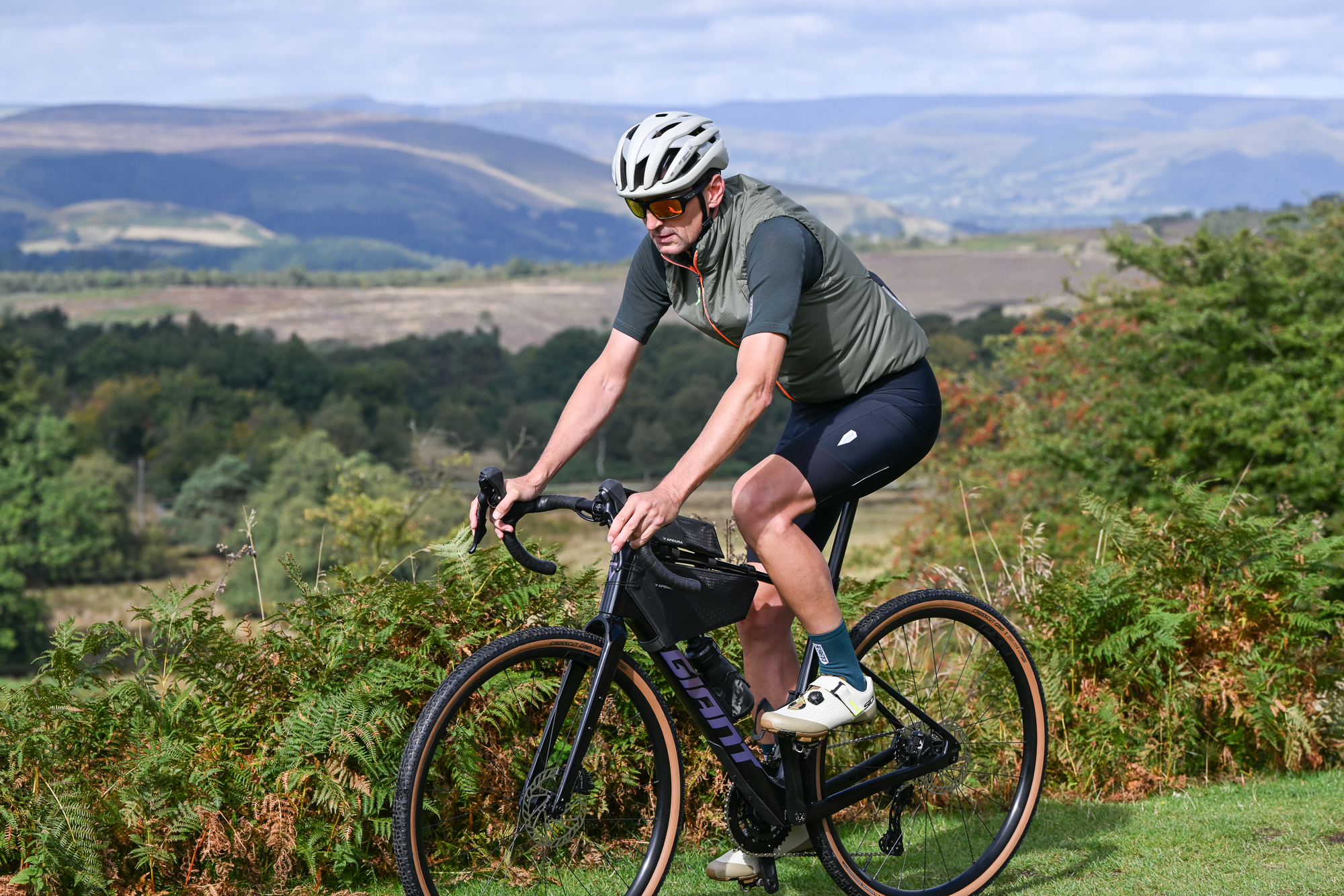 a male rider cycling a black gravel bike on a grassy trail wearing a green jersey, green gilet, black shorts and sunglasses