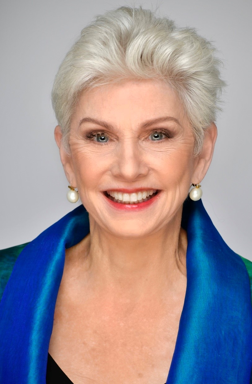 A headshot of a woman with cropped gray hair wearing a blue jacket smiling at the camera