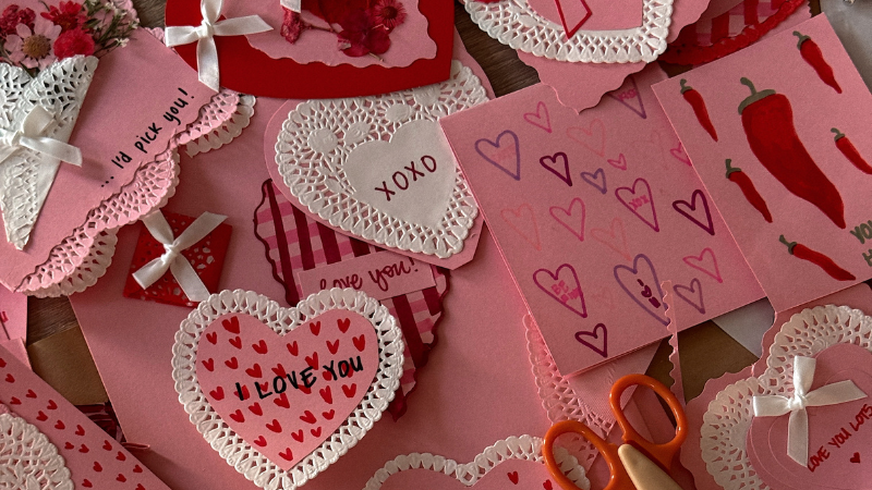  handmade valentine’s day cards in pink and red, decorated with lace, bows, and hearts, scattered with crafting supplies. 