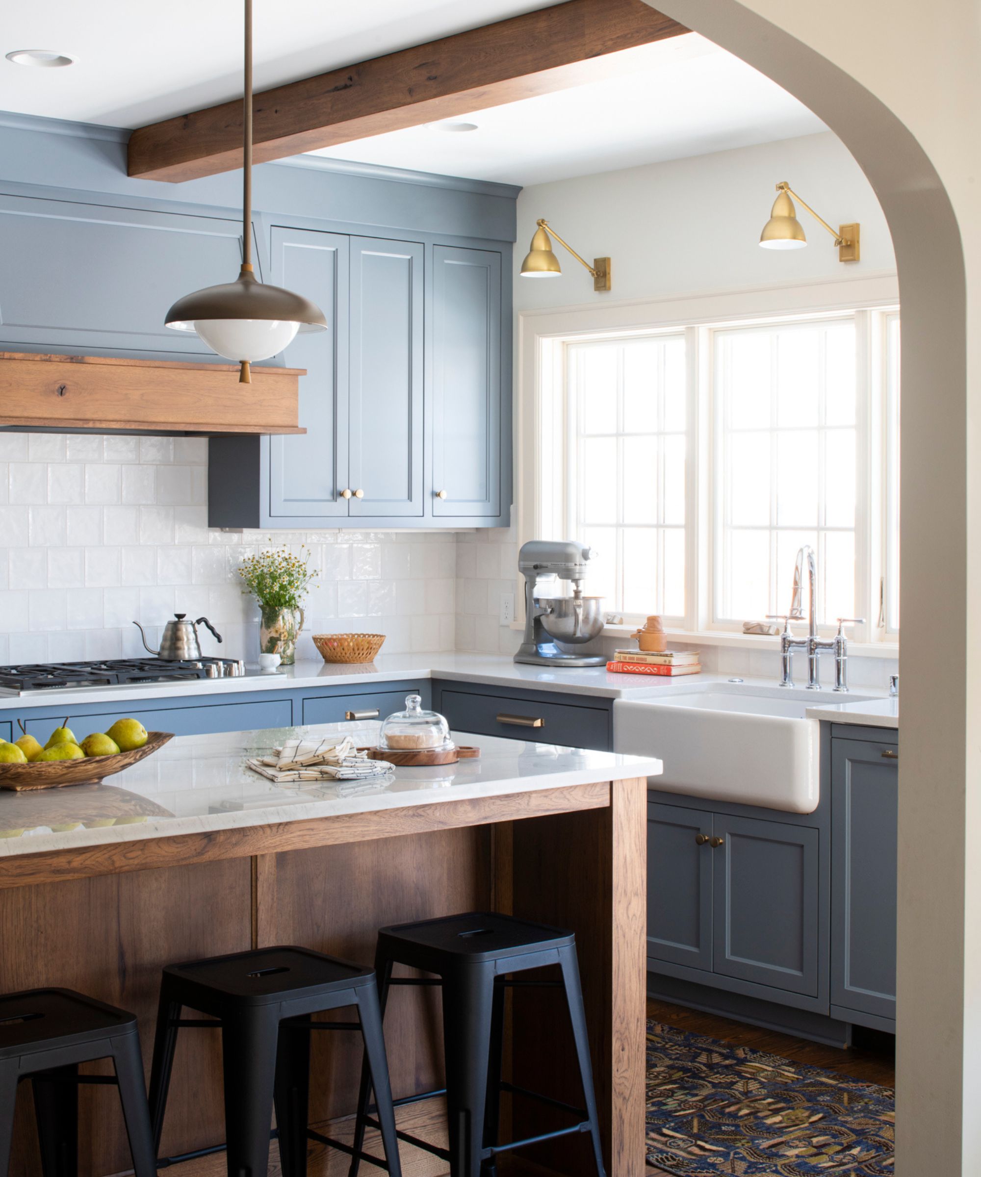 A transitional kitchen with blue-gray cabinets, a wooden island with white countertops, black bar stools, and wood ceiling beams. Brass wall sconces by the window and a butler's sink.