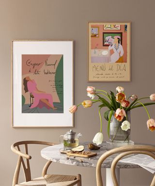 A boho dining room with a light brown wall with two wall art prints on it, and a gray marble dining table with a vase of tulips and two curved Scandi style wooden chairs in front of it
