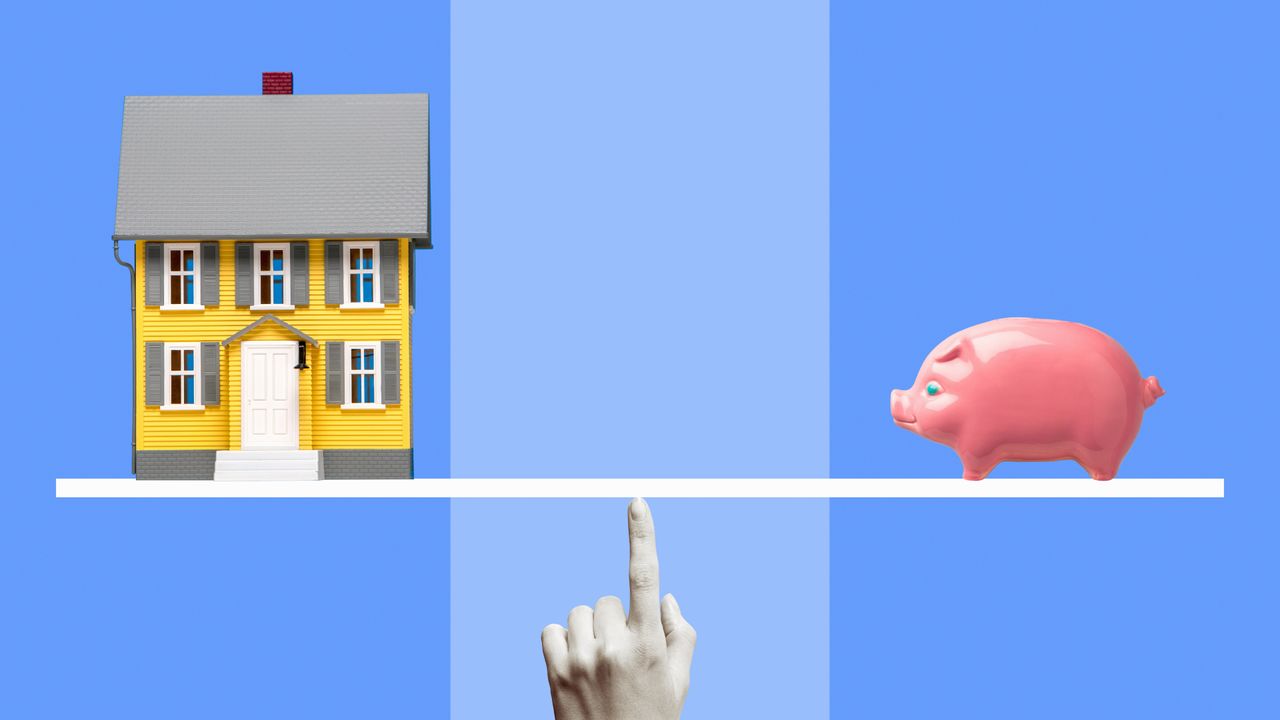 Yellow miniature model house and pink ceramic piggy bank on white line balanced on black and white woman&#039;s finger, blue background