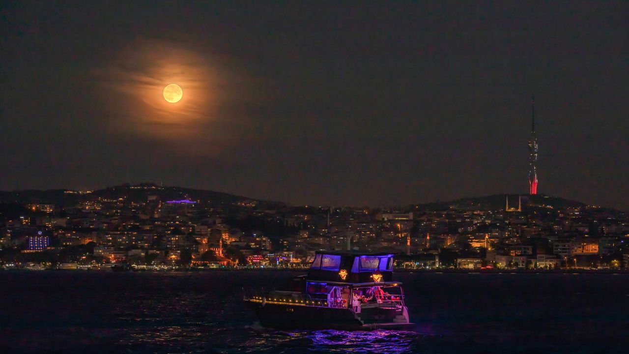 The cruise boat passing through the Bosphorus seen with the full moon appearing in the sky during the evening hours and the Camlica Mosque.