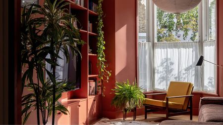 Minimal living room with earthy red painted walls and a built-in bookcase with TV, a lantern pendant light, a yellow midcentury chair, a brown boxy sofa, a Berber rug and a gold floor lamp in the corner beside a large bay window with cafe curtains and lots of large potted plants