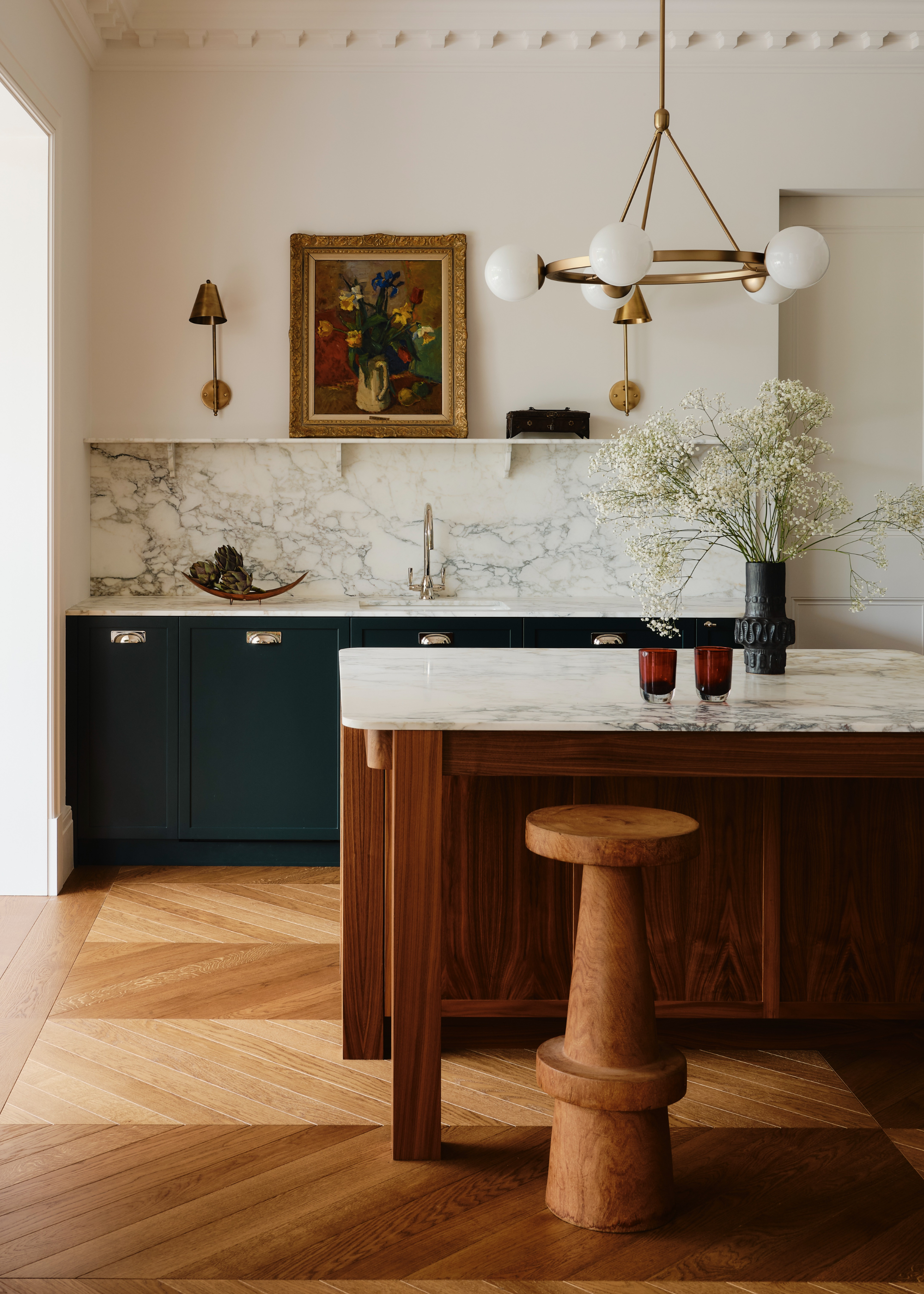 A west london kitchen with a dark wood island and a marble top, dark green cabinetry and a marble backsplash. Above the island is a gold light fixture