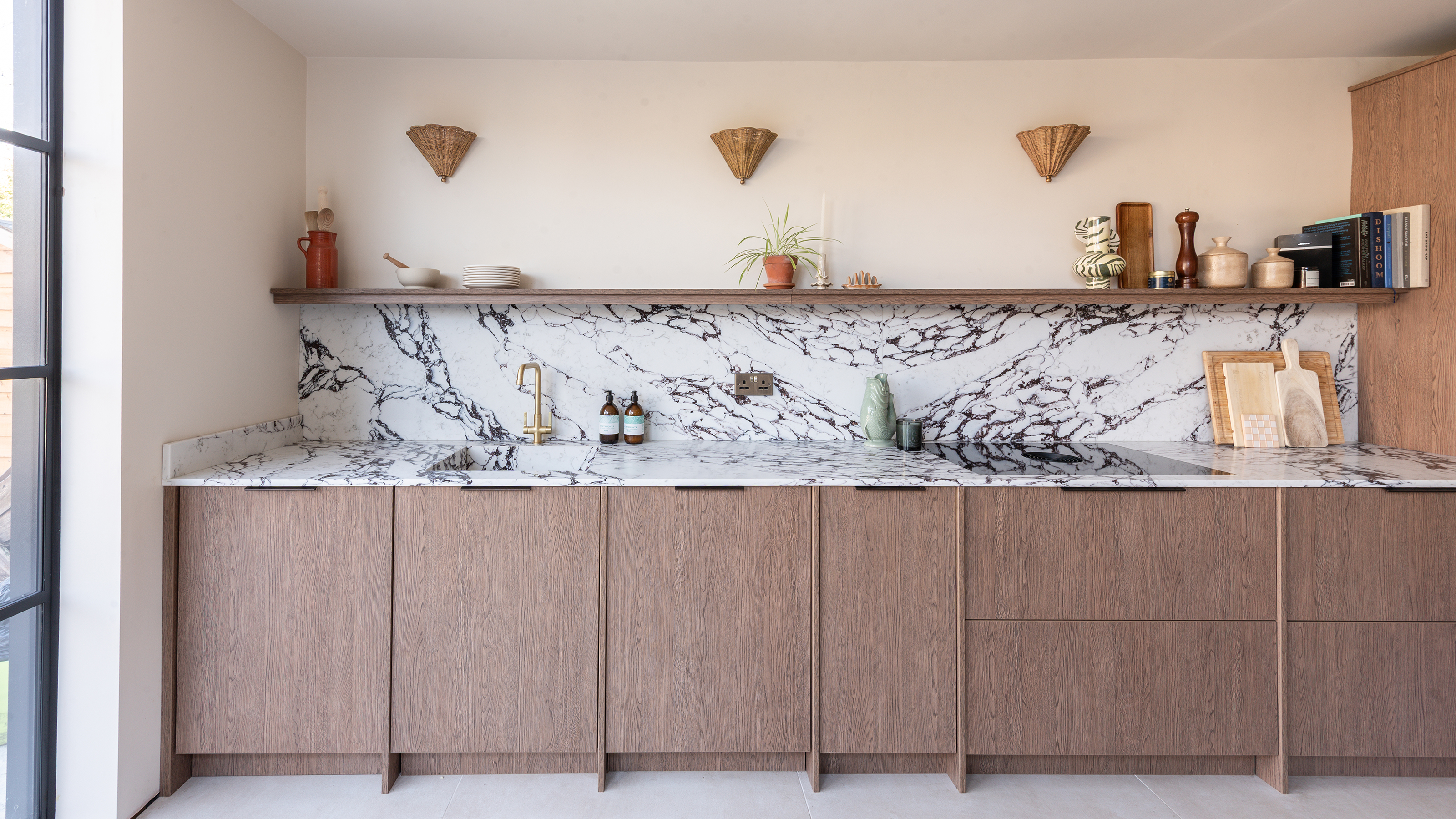 Single run of wood kitchen cabinets with a black and white marble worktop and splashback going up to an open shelf with three wall lights above it