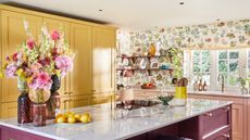  Colorful kitchen ideas are so fab. Here is a kitchen with tall yellow cabinets, a kitchen island with a vase of flowers, lemons, and utensils on top and a purple base, floral wallpaper and light pink cabinets