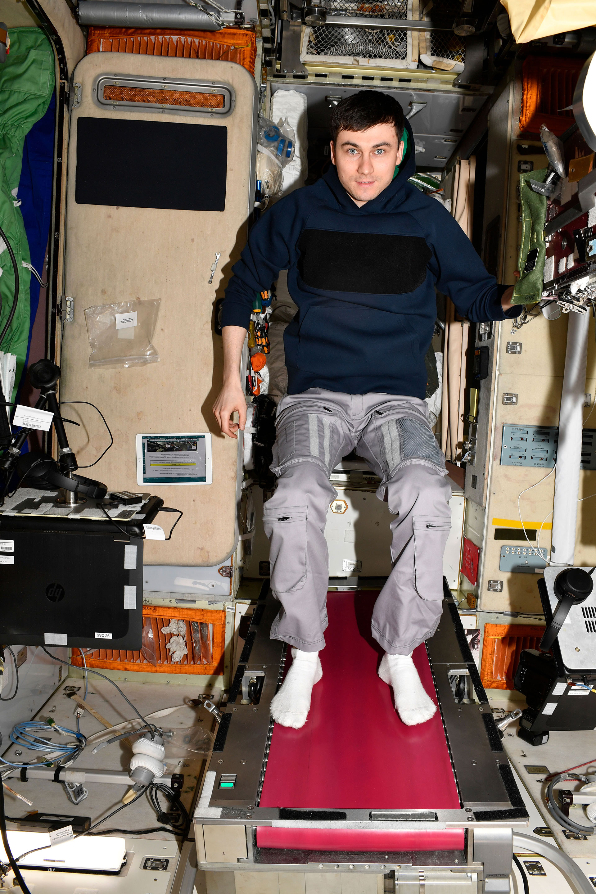 a man in a dark blue shirt and gray slacks stands atop a treadmill with red tread panels aboard a space station