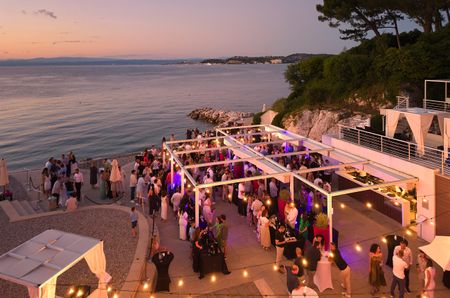 Birds-eye view of a drinks reception on the coast at sunset, with fairy lights above groups of people socialising