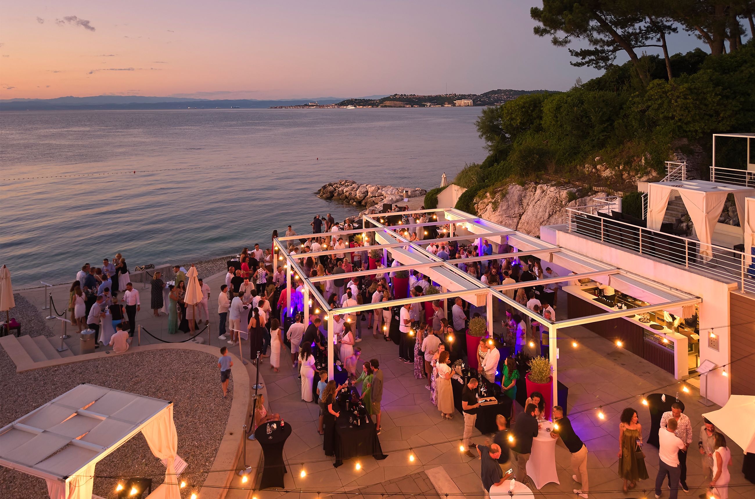Birds-eye view of a drinks reception on the coast at sunset, with fairy lights above groups of people socialising