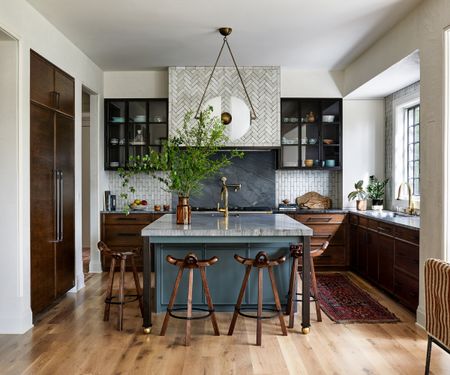 A large kitchen with dark wood cabinets, a mid blue island, marble countertops, and glazed wall cupboards