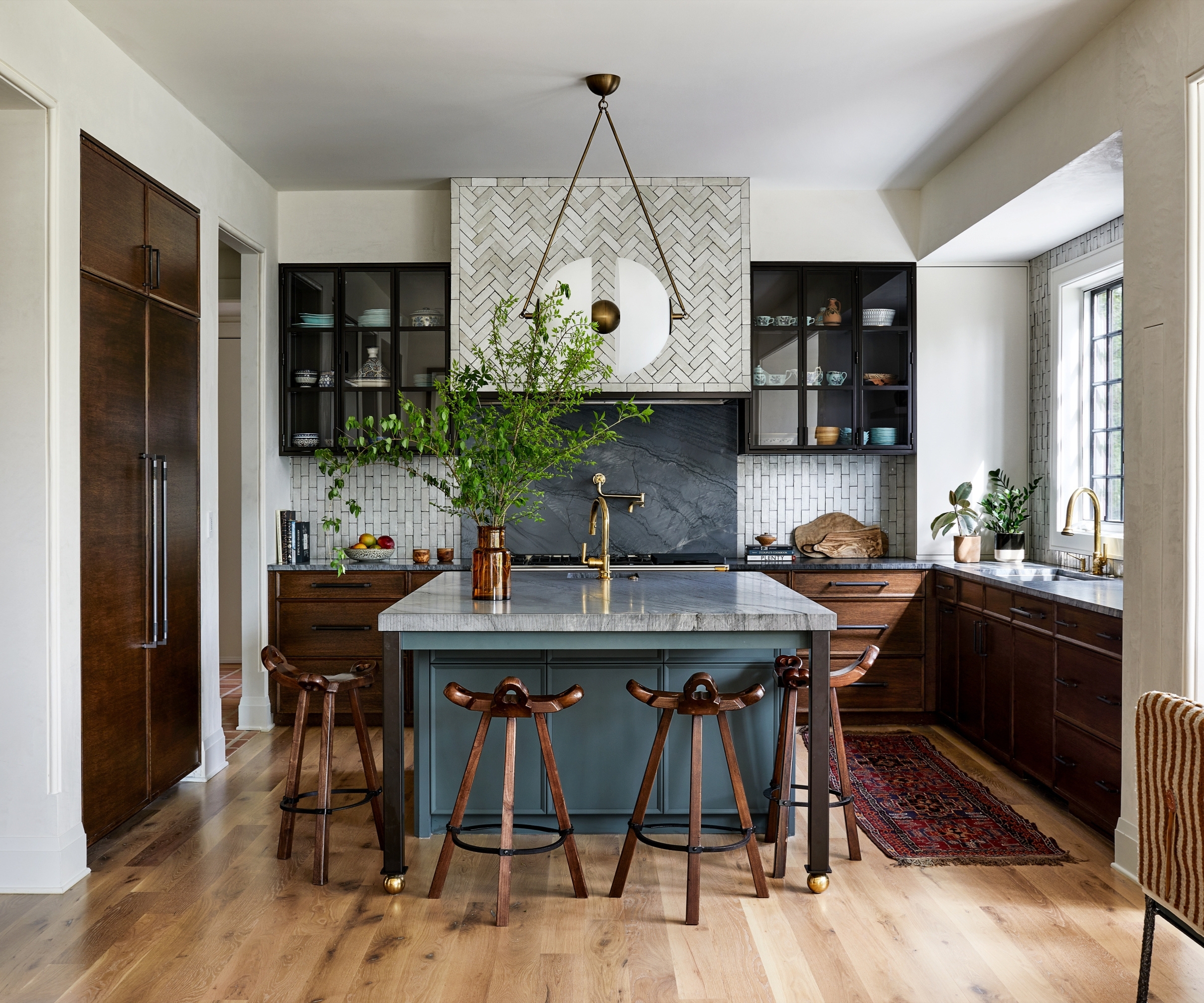 A large kitchen with dark wood cabinets, a mid blue island, marble countertops, and glazed wall cupboards