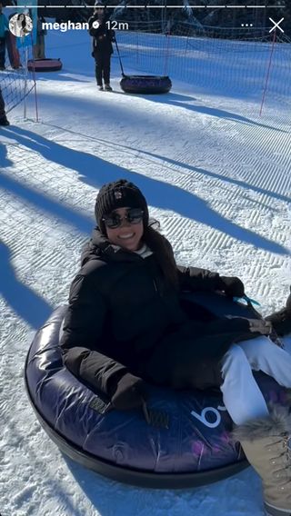 Meghan Markle wearing a black coat and hat and fur trimmed boots sitting in a snow tube and smiling