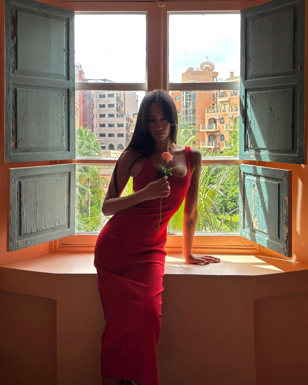 Content creator Emna Sellimi posing in a window wearing a red dress and holding a rose stem.