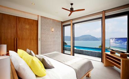 The Habita hotel room with floor to ceiling windows overlooking ocean, white bedding, yellow cushions and wooden TV unit