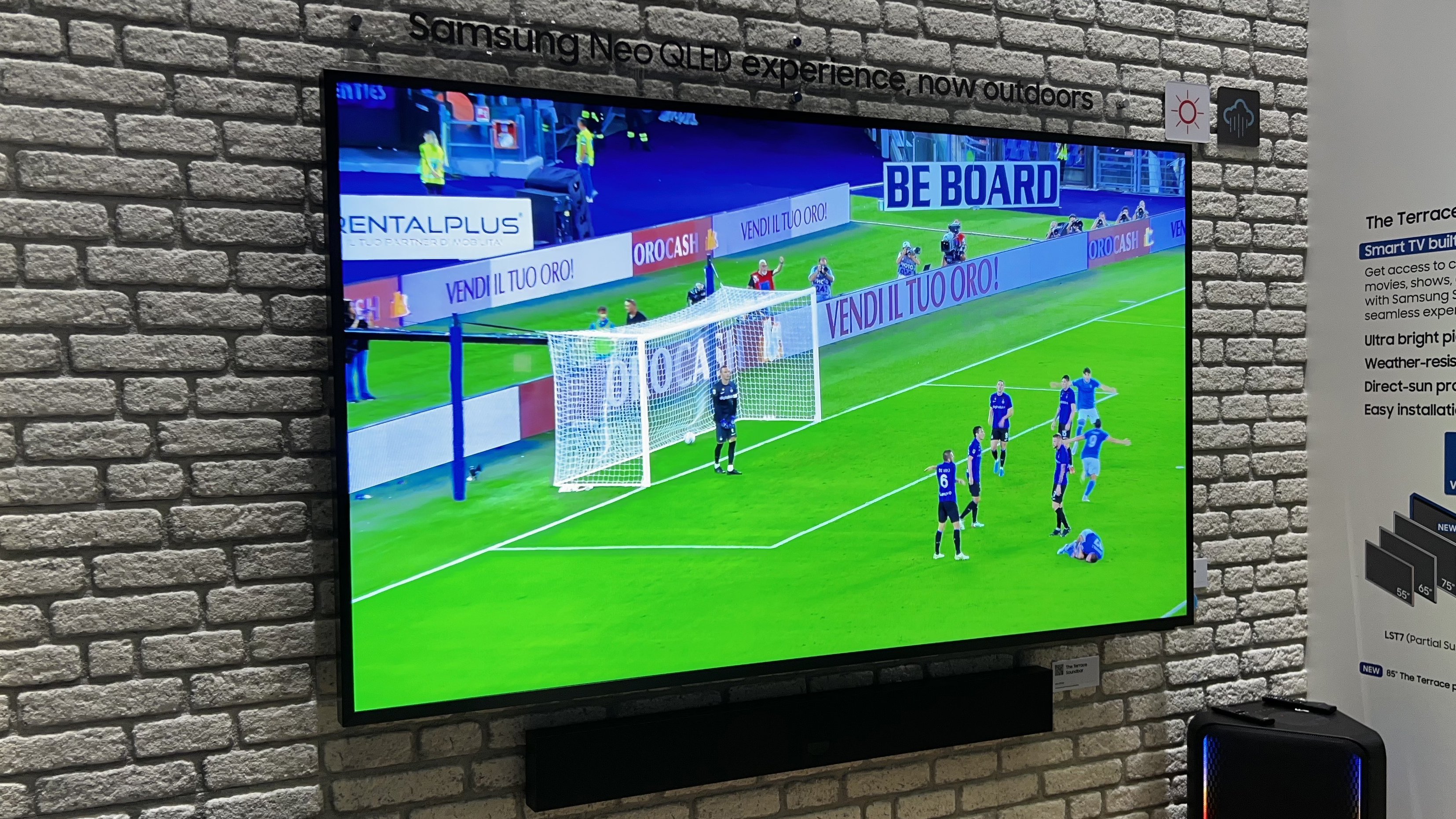 The Samsung Terrace TV on a brick-style wall, with a football match on the screen
