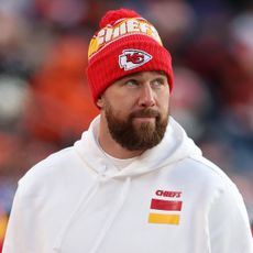 Travis Kelce wearing a red knit Chiefs hat and white Chiefs hoodie looking up to the left and smirking