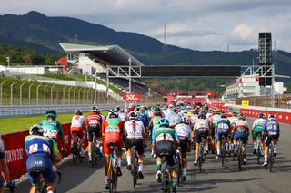 This is an image of the backs of riders racing at the Olympic 2020 showing all their race numbers and colourful kit. In the background are green hills and the edges of the race track that the riders are on.