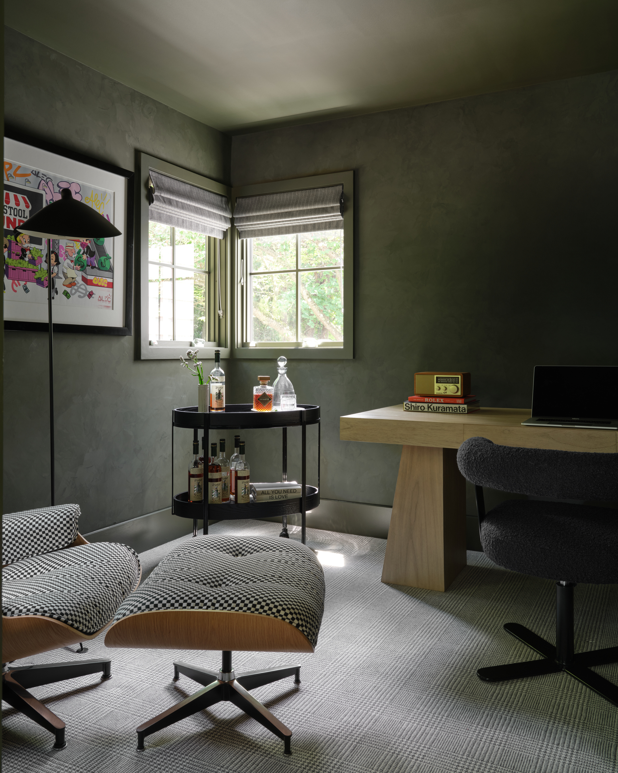 Image of a darker living room/home office painted in an olive green. There is a large wooden desk, a black bar cart, and two fabric chairs.
