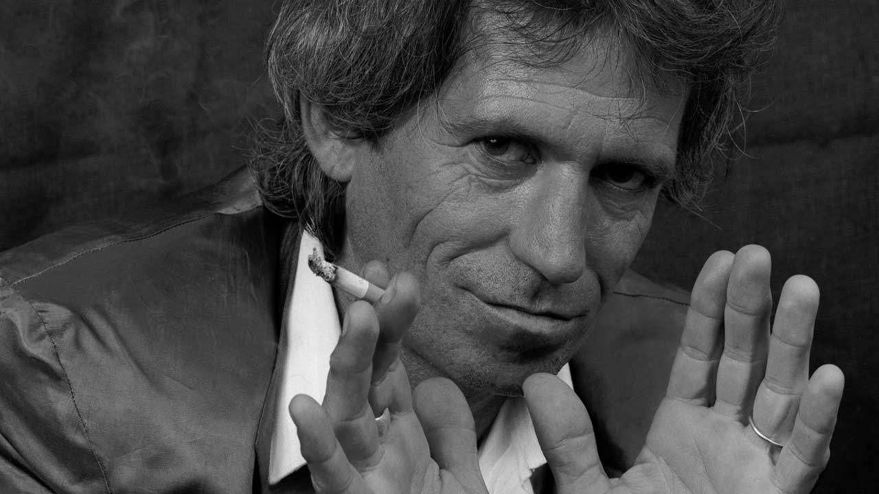 Keith Richards in 1987, smoking a cigarette and holding his hands up towards the camera