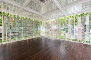 The interior of a pavilion with a dark wood flooring and white square wire walls looking onto green grass.