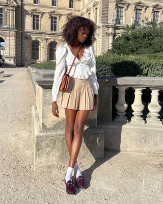 Woman on street wears cream boho blouse, beige pleated skirt and loafers