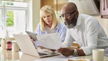 An older couple look at paperwork together while sitting in front of a laptop at their kitchen table.