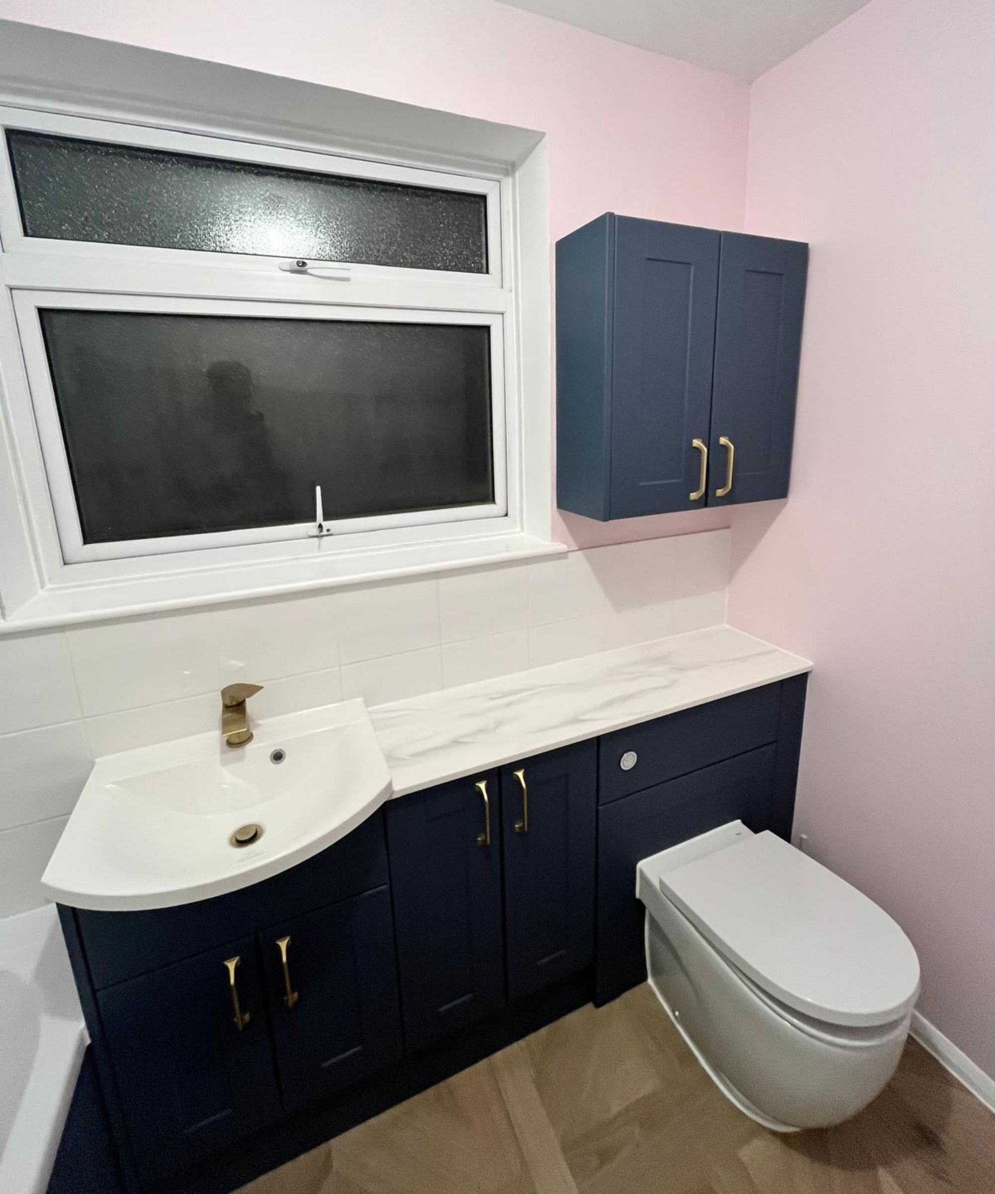 Renovated bathroom with navy blue shaker-style fitted units, gold brass taps and handles, marble-effect worktop, white metro tiles and soft pink walls.