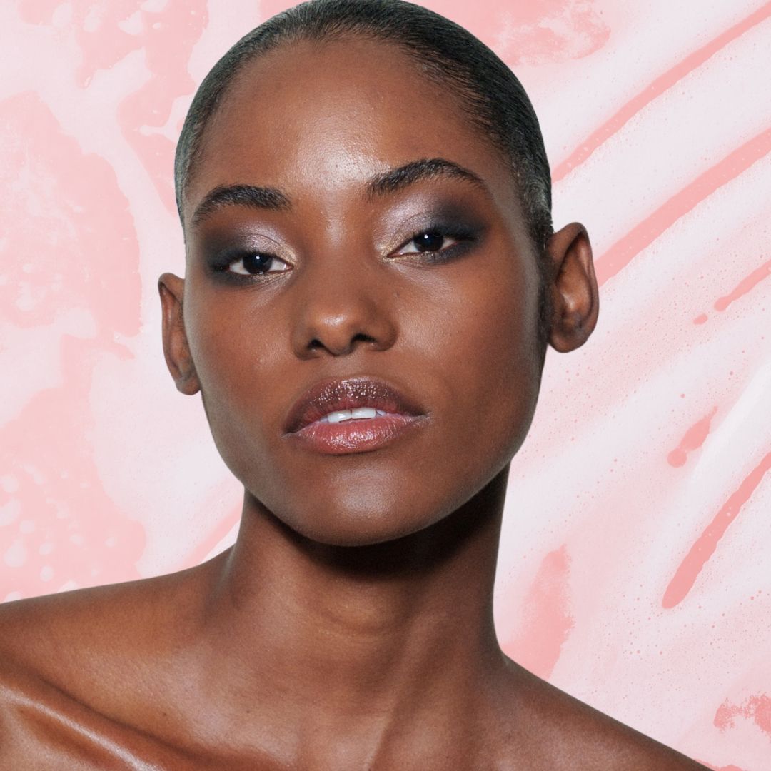 A woman with glowing skin wearing an off the shoulder top in front of a pink background that has soap suds on it. She in next to three beauty products from Brazilian brands.