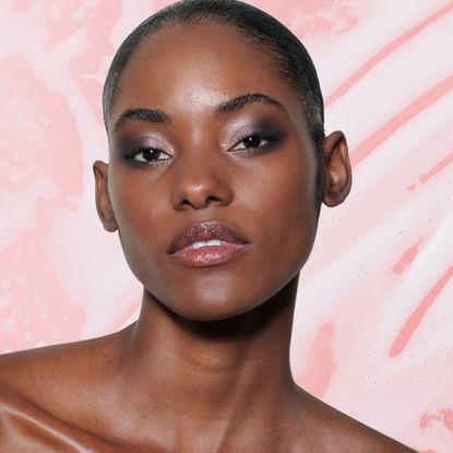 A woman with glowing skin wearing an off the shoulder top in front of a pink background that has soap suds on it. She in next to three beauty products from Brazilian brands.