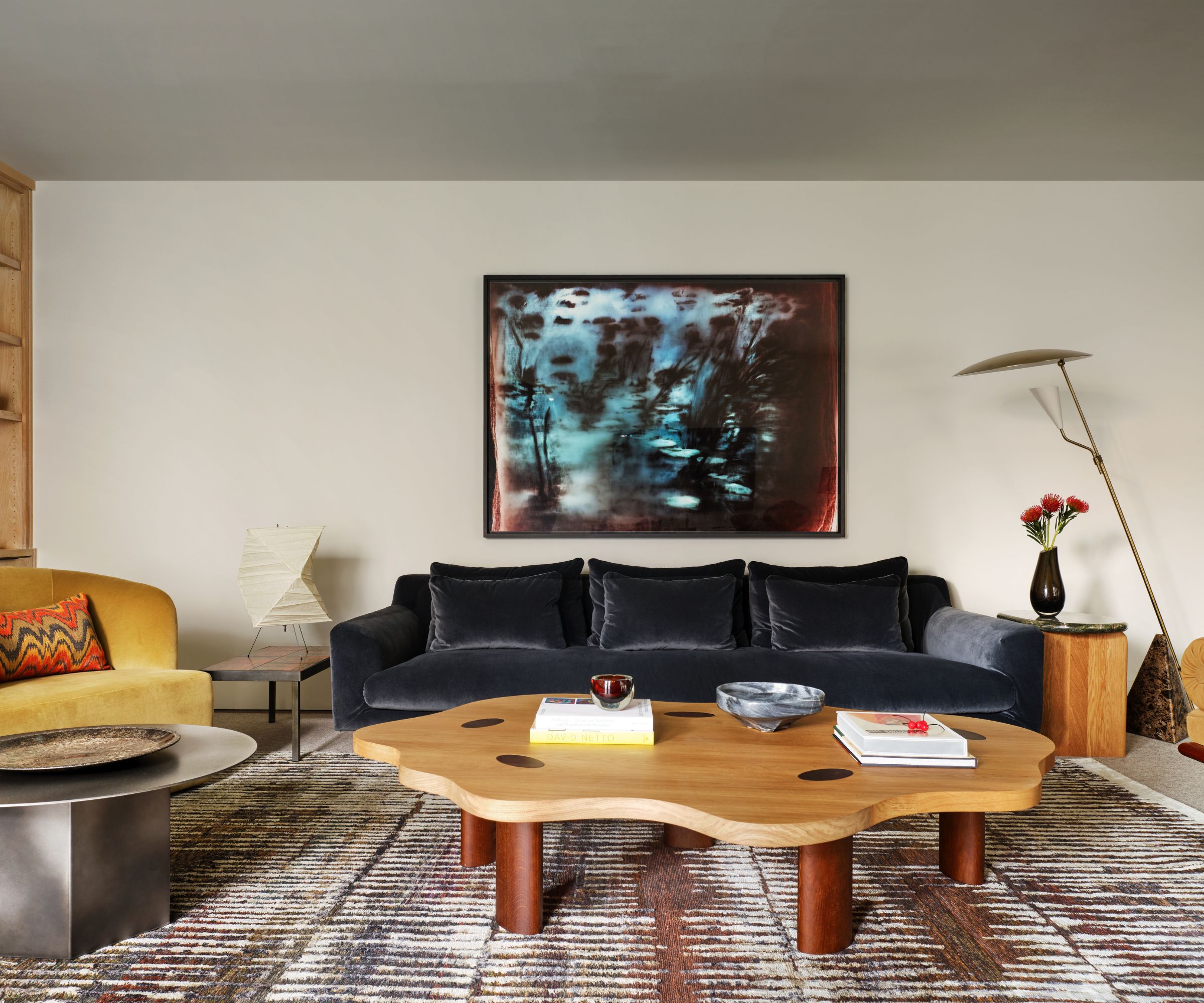 Living room with blue velvet sofa, curved wood coffee table, yellow armchair, patterned rug, floor lamp and abstract art