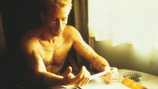 Guy Pearce as Leonard Shelby covered in tattoos and reading a note at a table shirtless in the thriller movie Memento.