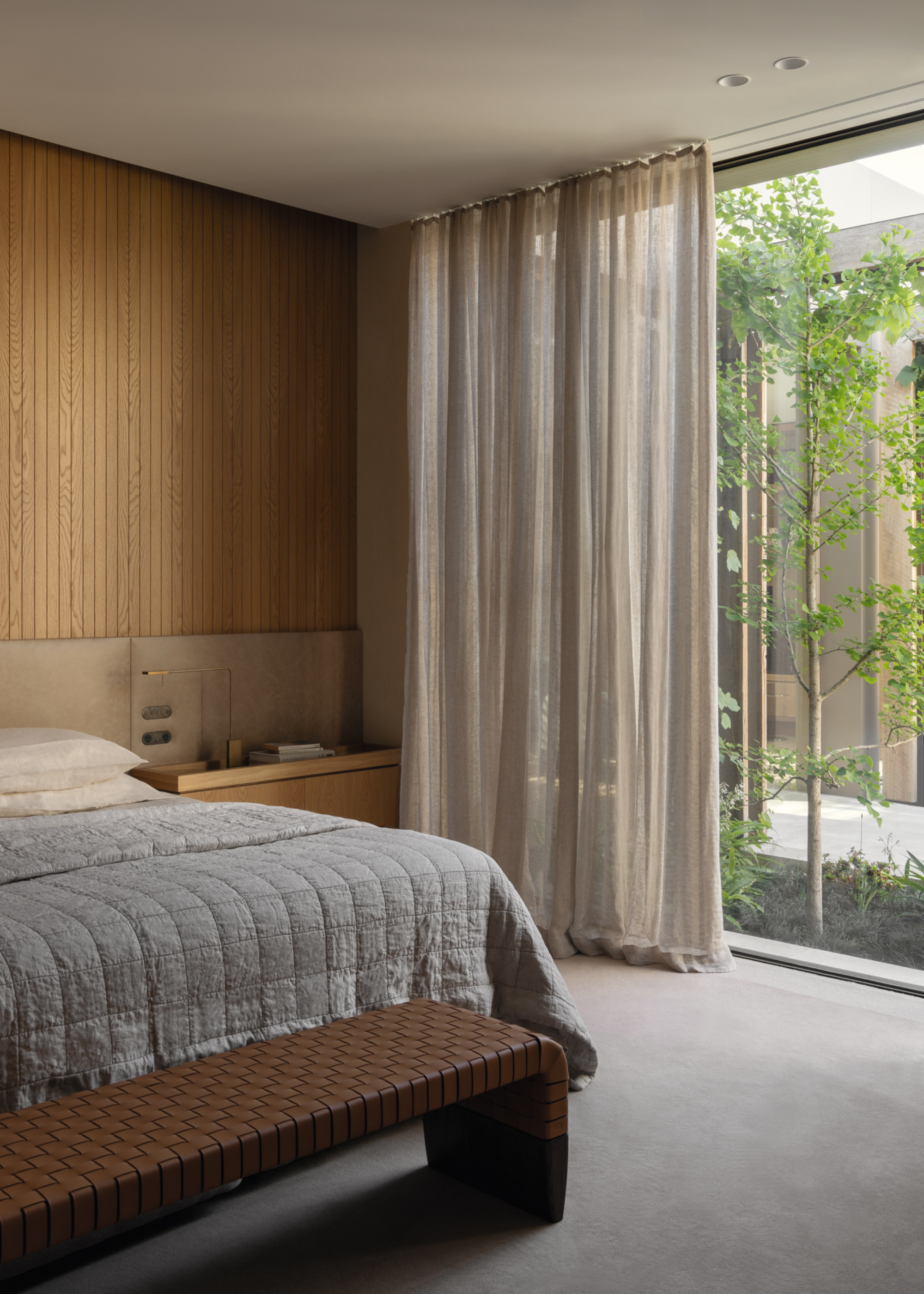 A bedroom with large windows, beige sheer curtains, a leather bench, and a simple bedscape