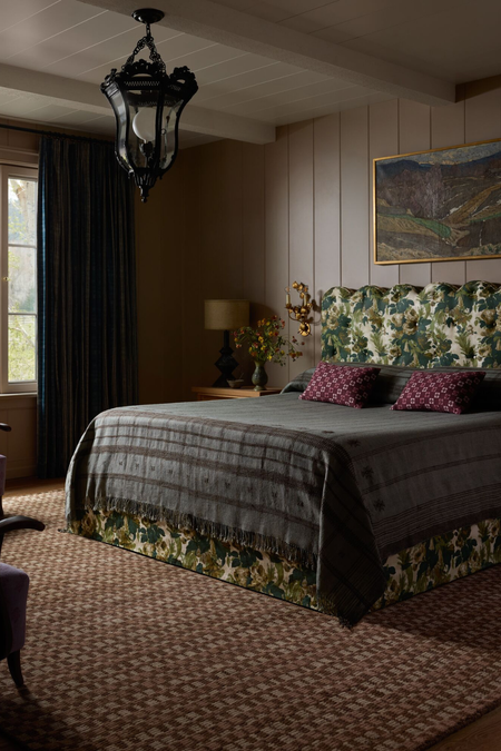 Moody, richly layered bedroom featuring a neutral heritage patterned rug, tapestry-print bed frame flanked by two ornately gilded candle sconces, and eclectic bedding.