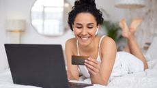 A woman with curly dark hair wearing a white nightshirt lies on her stomach, smiling, while holding a bank card and shopping for an affordable mattress online