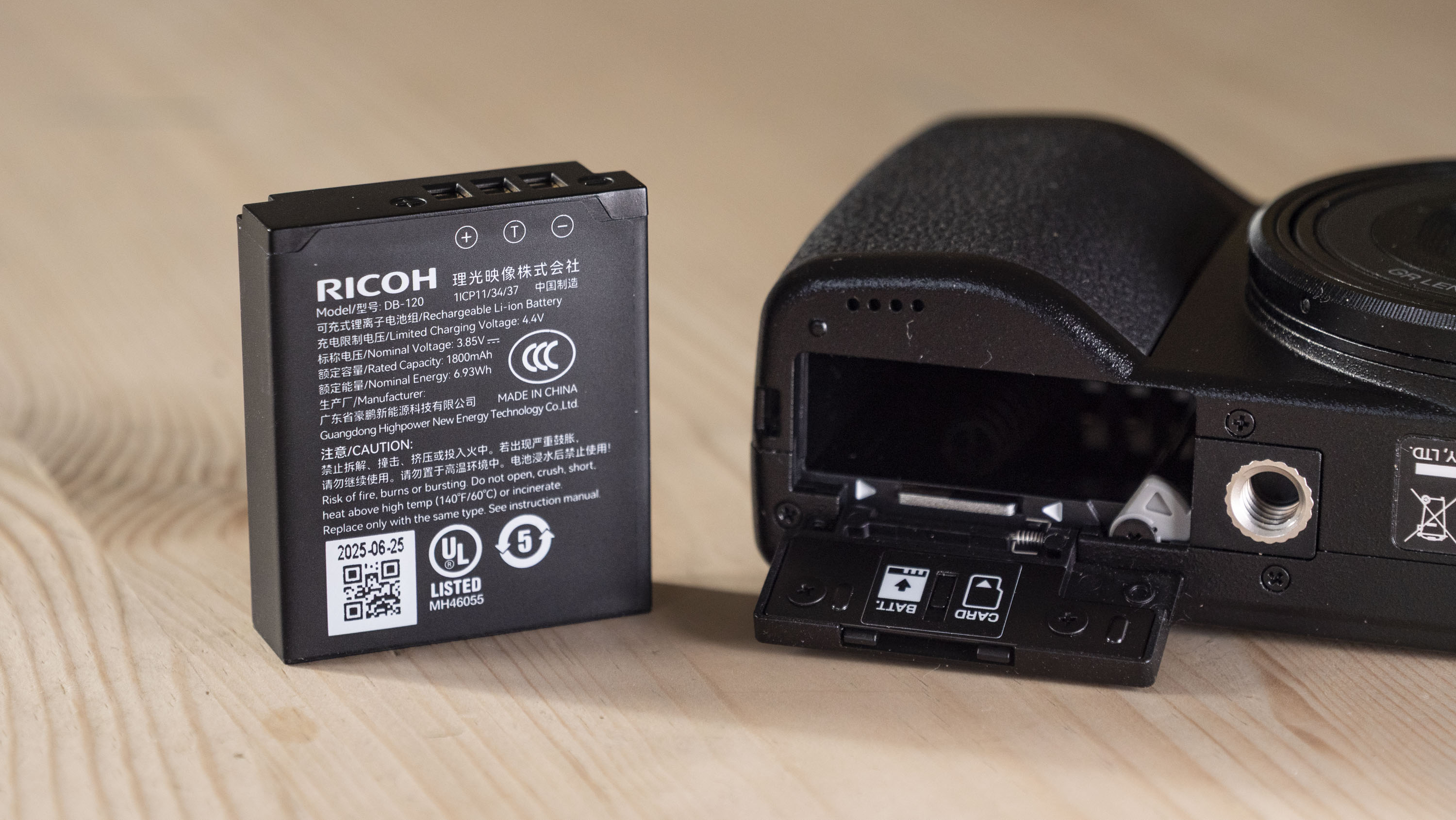 Ricoh GR IV compact camera on a wooden table, alongside its battery