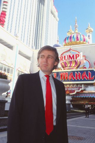 Donald Trump wearing a red tie and blue suit standing outside the Trump Taj Mahal in 1990