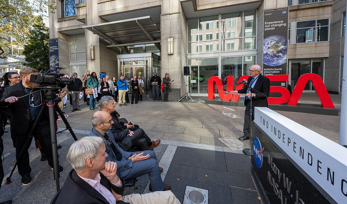 a man in a blue suit speak in an outdoor courtyard with &quot;nasa&quot; written in red in the background.