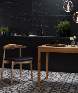 A black kitchen with black wall tiles and a countertop with a black kitchen faucet, a warm wooden table and a wooden chair with black cushioning, and white and gray tiling