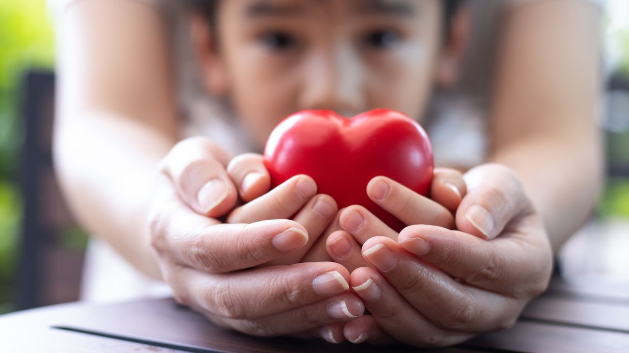 An adult cups her hands around the hands of a child who&#039;s holding a heart knickknack.