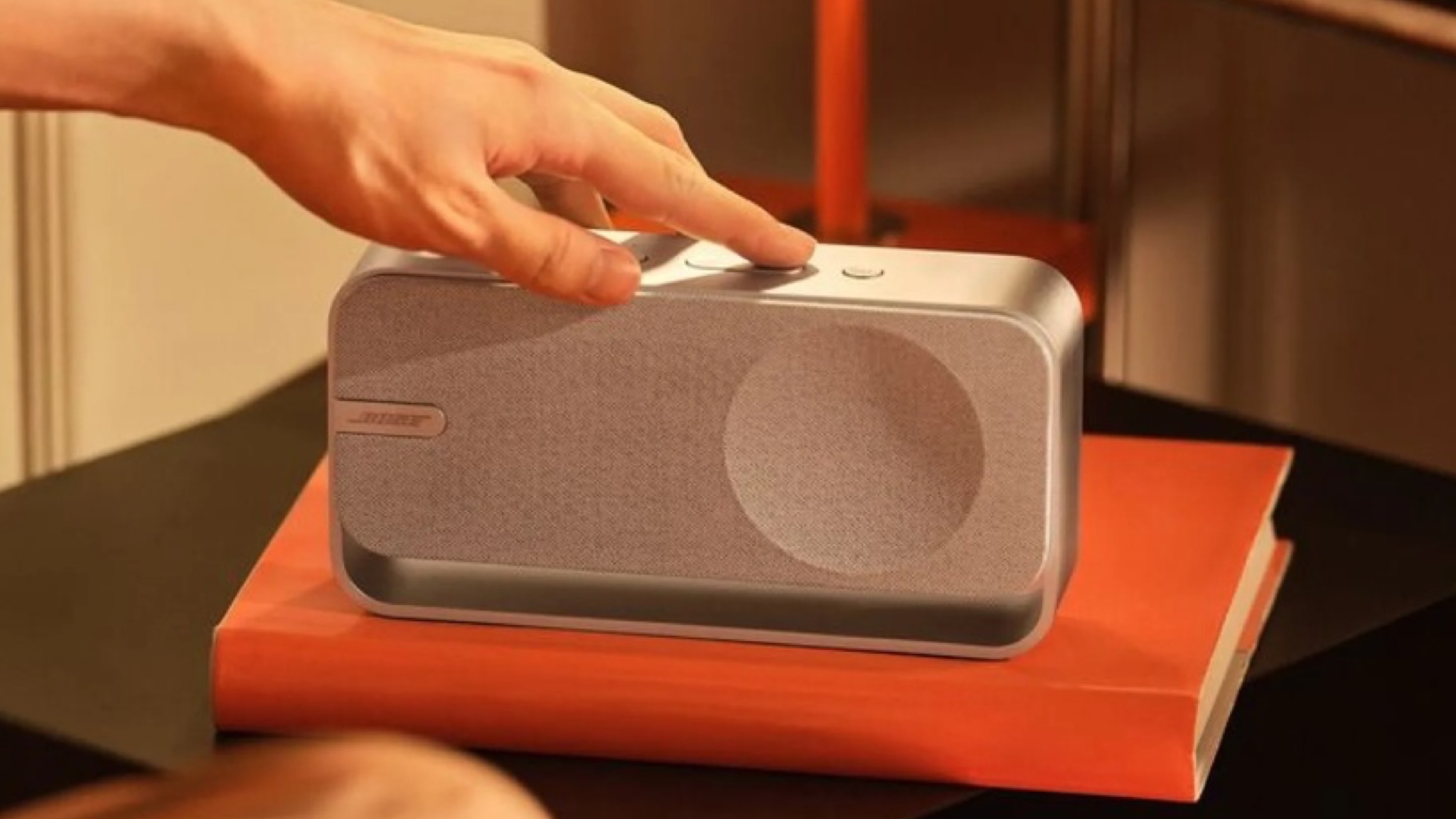 Bose SoundLink Home speaker with a person's hand pressing one of the buttons on top. The speaker is sat on an orange book, which in turn is on a black surface