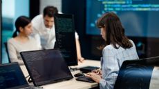 Female software developer with long dark hair, wearing a striped suit jacket, coding on a desktop computer with multiple screens while co-workers confer in background.