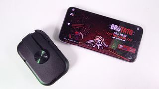 Folded, compact Razer Prio mobile controller resting next to a smartphone displaying the main menu of the game Brotato on a white surface.
