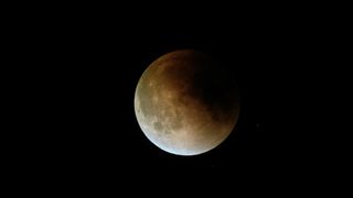 lunar eclipse with a dark red shadow in the upper right of the moon and a light white color at the bottom left.