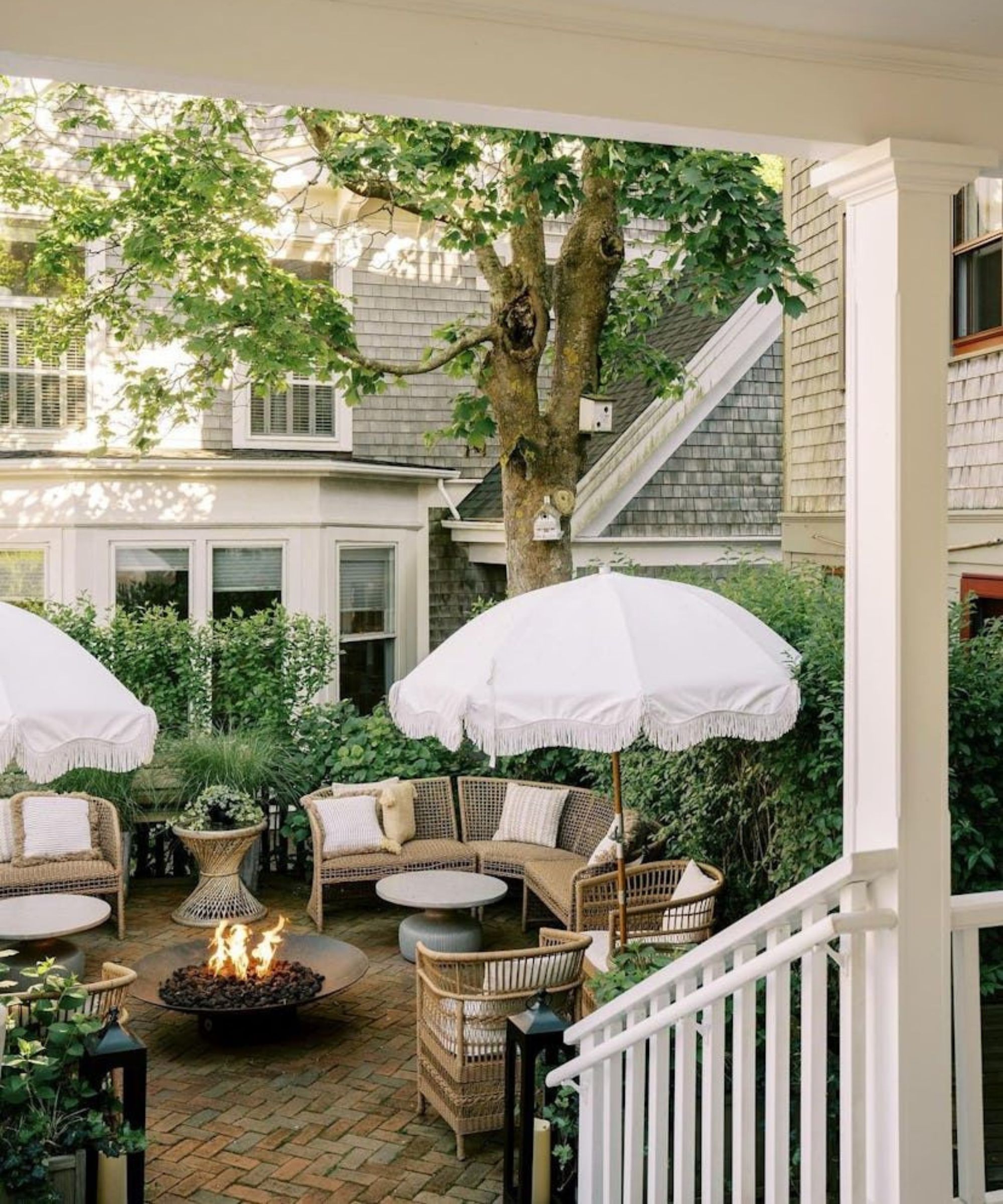 small hamptons style outdoor backyard with stairs down to a brick paved patio surrounded by greenery with wicker furniture and white fringed patio umbrellas