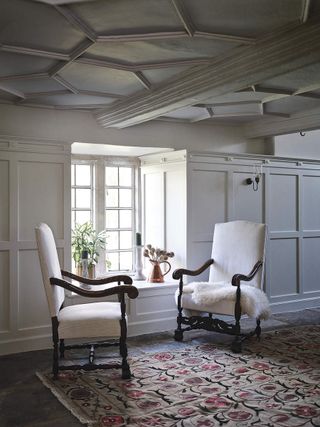 Panelled walls and ceiling, window with deep sill. Period wooden chairs upholstered in cream with faux fur throw and floral rug on stone floor.